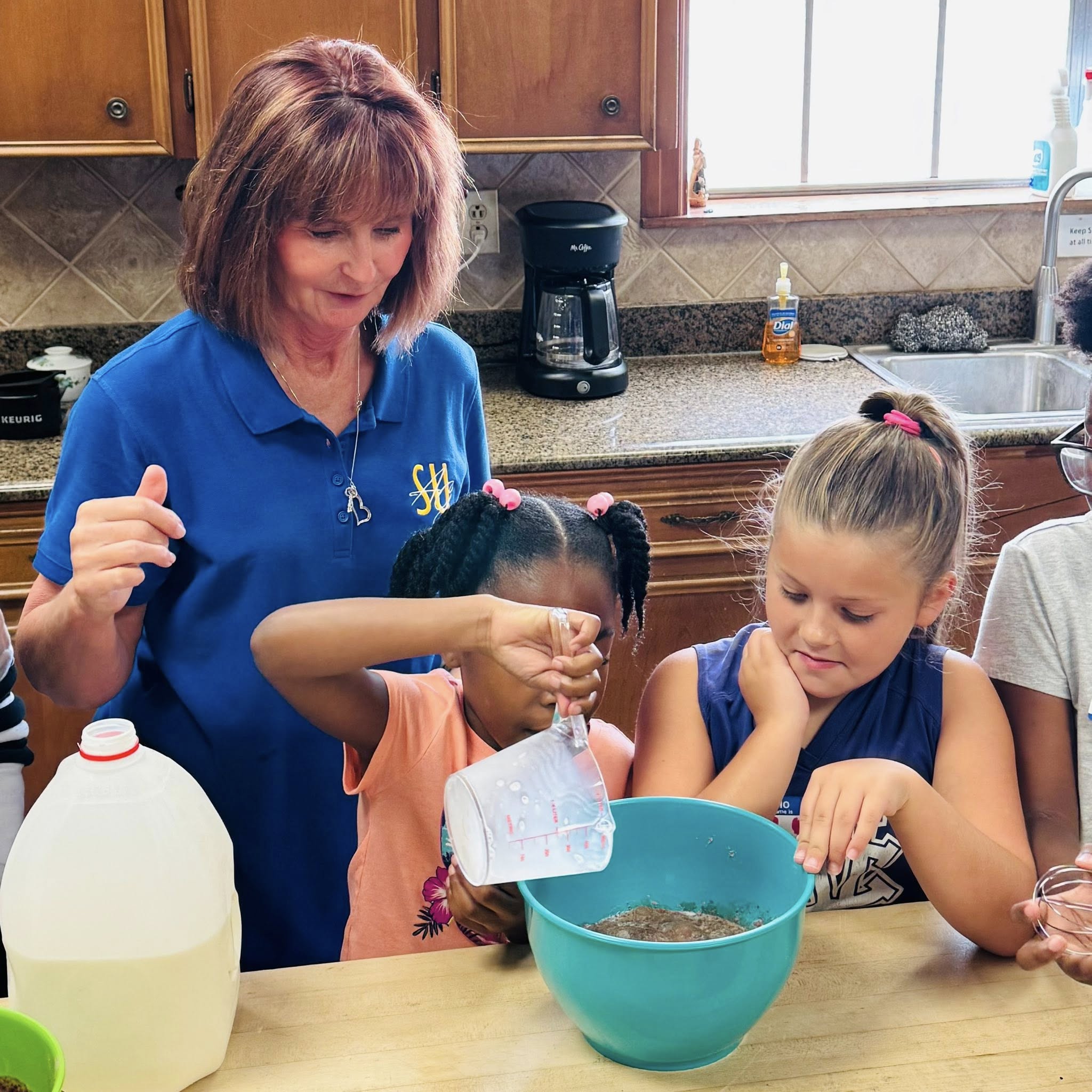A woman and a girl watching another girl pour something in a bowl.